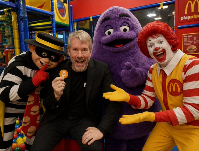 A man holding a Bitcoin coin poses with the Hamburglar, Grimace, and Ronald McDonald at a McDonald's PlayPlace, with a man resembling MicroStrategy's Michael Saylor in the center.