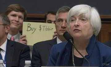 A woman with white hair speaks at a microphone during a hearing, while a man behind her holds up a sign reading 'Buy $Tibbir'.