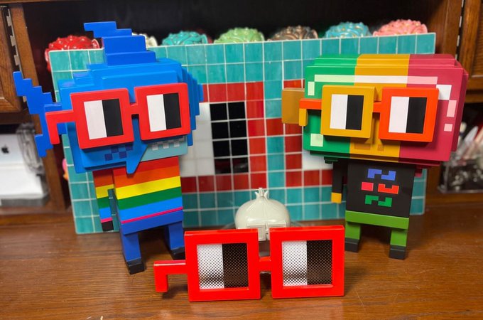 Two pixel-art style figures with 3D glasses—one blue with a rainbow shirt and one green and yellow with a pixel face—stand before a tiled background with red 3D glasses in the foreground.