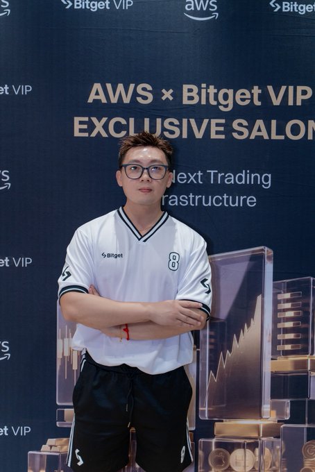 A young man wearing a white Bitget #8 jersey and black glasses stands with arms crossed in front of a backdrop reading "AWS × Bitget VIP EXCLUSIVE SALON." The branded Bitget apparel and event setting visually reinforce his role as a BG TradFi (Traditional Finance) spokesperson for the cryptocurrency exchange.