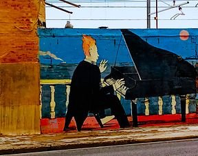 Street art mural depicting a cartoon character with spiky blond hair in a suit playing a grand piano against a colorful blue and orange wall.
