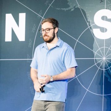 A man in a blue polo shirt and glasses speaking on stage in front of a world map backdrop with partial large text visible.
