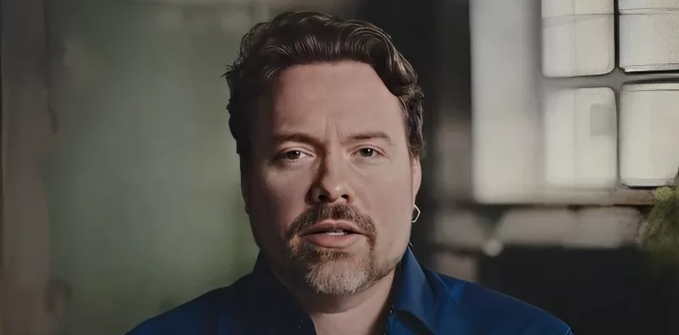 A man with short brown hair and a goatee, wearing a blue shirt and an earring, speaks directly to the camera in what appears to be an interview setting.