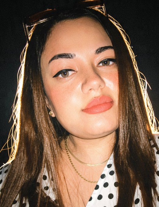 A close-up portrait of a young woman with long brown hair, sunglasses on her head, winged eyeliner, and orange lipstick, wearing a polka dot top and gold necklaces.