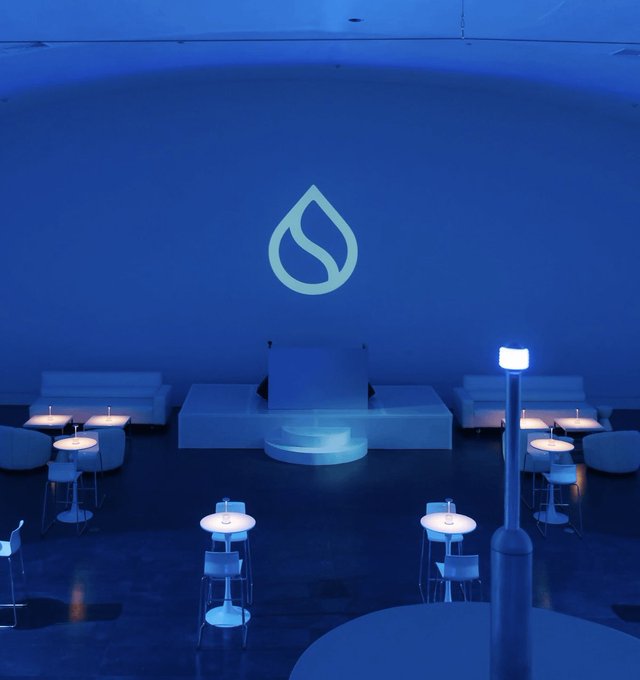 A dimly lit, blue-toned room featuring a large illuminated water drop logo on the wall above a white seating area, with several small round tables and modern stools arranged in the foreground.