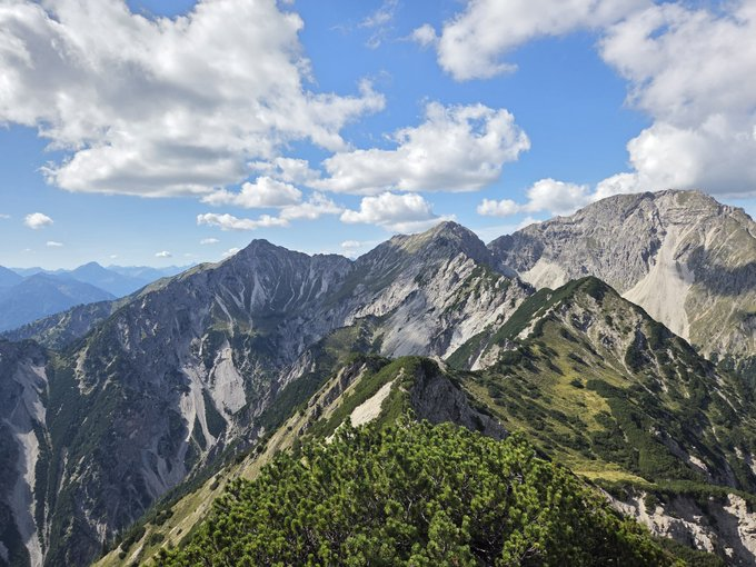 A scenic mountain landscape featuring rugged gray peaks, green vegetation in the foreground, and a blue sky filled with scattered white clouds.