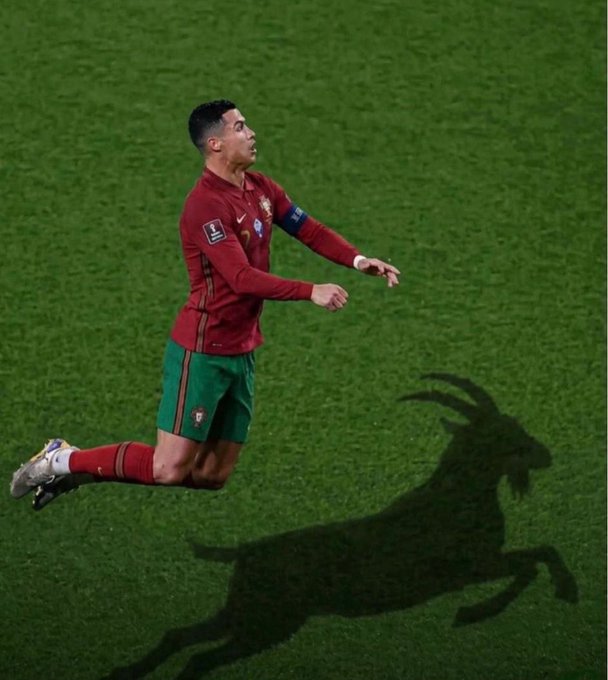 Portugal's Cristiano Ronaldo, wearing the captain's armband, runs across the pitch in his red and green national team kit while his leaping shadow is cast as a goat on the grass—a nod to his iconic "GOAT" (Greatest of All Time) status.