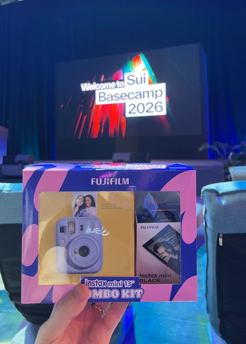 A hand holding a Fujifilm instax mini 13 CAMEO KIT box (lavender colored camera) in front of a stage screen displaying 'Welcome to Sui Basecamp 2026' with red brushstroke graphics.