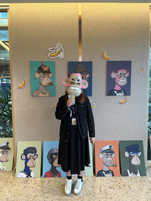 A person standing in front of an art display featuring cartoon monkey NFT-style paintings and banana stickers, holding a drink.