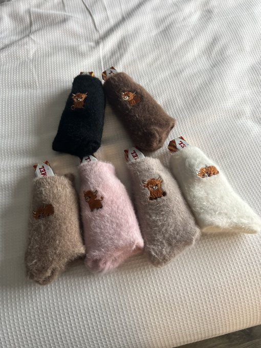 A photo showing six fuzzy, slipper-style socks in various colors—navy, brown, tan, pink, gray, and white—each featuring a small Highland cow embroidered on the front, arranged on a white textured surface.