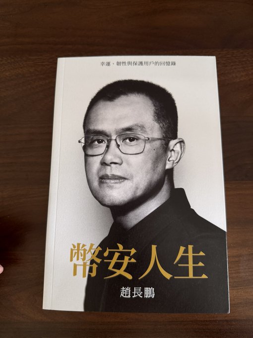 A Chinese book titled "币安人生" (Binance Life) by 赵长鹏 (CZ, Changpeng Zhao), subtitled "幸运、耐性与保护用户的回馈" (Luck, Patience, and the Rewards of Protecting Users), rests on a wooden table. The post celebrates finally obtaining a physical copy of CZ's book about his Binance journey.