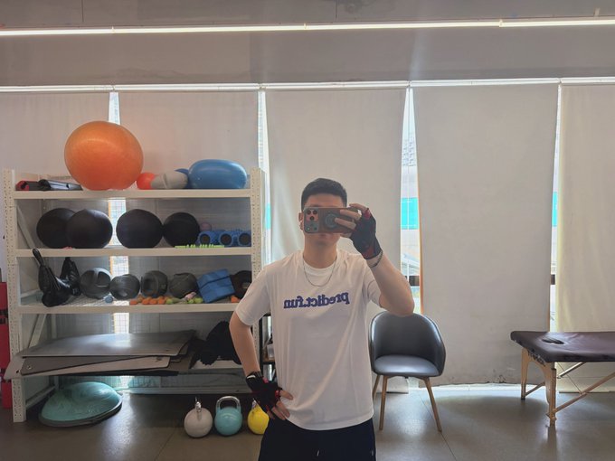 The image features a man taking a selfie with his cell phone, standing in front of a gym filled with exercise equipment such as dumbbells and kettlebells. The gym appears to be well-equipped for various workouts, including weightlifting and cardio exercises. The man is wearing a white t-shirt that says "Prediction" on it, which adds a unique touch to the photo.