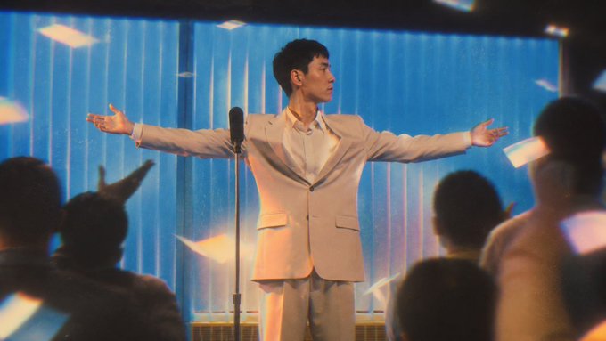 A man in a light-colored suit stands on stage with arms outstretched before a microphone, surrounded by an audience amid falling papers, evoking a celebratory or performance moment. The image's triumphant atmosphere aligns with the post's promotion of a newly launched, verified NFT mint collection at a discounted rate on OpenSea.