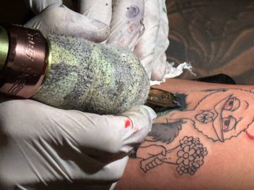A tattoo artist wearing gloves uses a vibrating tattoo machine to ink an outlined design featuring a woman's face, a wrapped candy, and grapes onto a client's skin. The image's graphic nature and underground aesthetic visually echo the post's themes of raw, back-end labor and dedication to community projects like meme curation and event hosting on platforms such as Canna and memedepot.