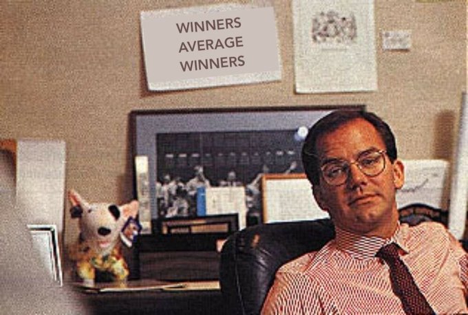 A man in glasses and a red tie sits in an office beneath a sign reading "WINNERS AVERAGE WINNERS," with a small chihuahua figurine beside him. The meme humorously frames the scam memecoin pump as the man embodying the smug, self-satisfied "average winner" despite the dubious nature of the project.