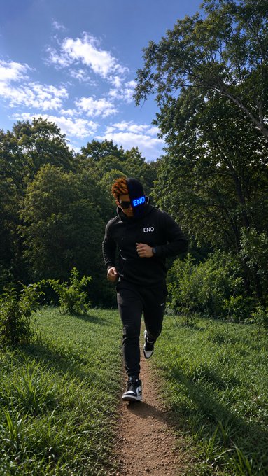 A person wearing a black 'END' hoodie and headband runs along a dirt trail through a grassy forest under a blue sky.