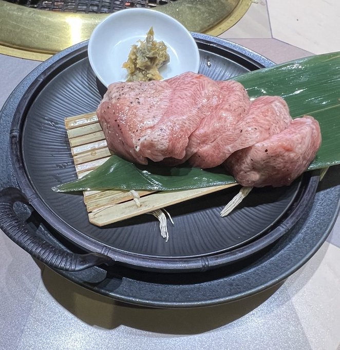 Raw marbled meat is presented on a dark plate over a green leaf and bamboo mat, accompanied by a small dish of seasoning, likely at a Korean barbecue restaurant.