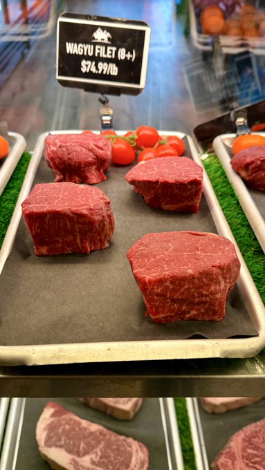 A display case in a butcher shop or market shows thick, raw cuts of red meat on a tray, labeled with a sign that reads 'WAGYU FILET (B+) $74.99/lb,' with cherry tomatoes on the side.