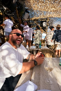 A bearded man in sunglasses laughs joyfully at an outdoor daytime gathering, seated at a wooden bar table with a glass of white wine in the foreground. The festive scene, with crowds mingling beneath draped lights and trees, captures the carefree celebratory mood referenced in the post's playful request to "rain on" him and his kids.
