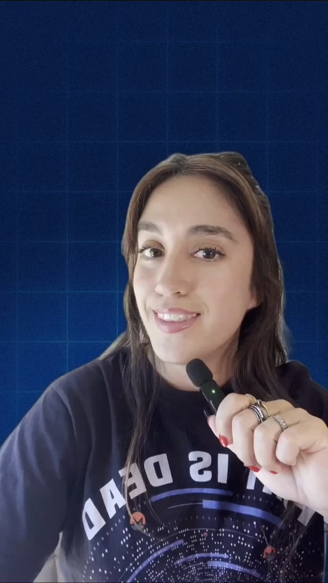 A smiling woman holding a lapel microphone, wearing a dark shirt with 'IS DEAD' partially visible in mirrored text.