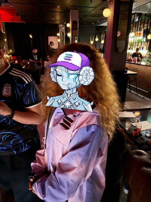 A person wearing a light purple jacket and carrying a pink backpack stands in a bar, with a pixel art sticker covering their face.