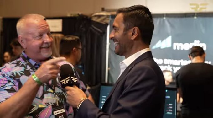 A reporter with an "RF" microphone interviews a smiling man in a suit at what appears to be a trade show, with a "martin" branded booth visible in the background—relating to the post's mention of a cameo appearance by Ray Fuentes during an on-camera segment.