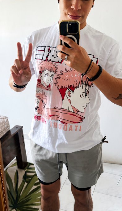 A mirror selfie of a person wearing a white t-shirt featuring an anime character with pink hair (Itadori Yuji from Jujutsu Kaisen) and Japanese text, paired with gray shorts, while making a peace sign.