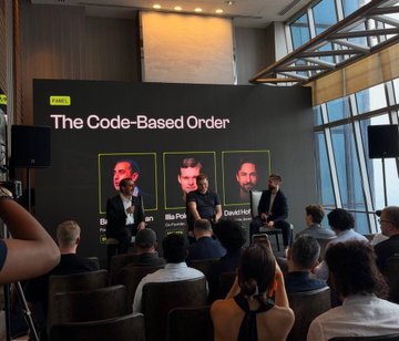 A panel discussion at a conference with three speakers seated on stage in front of a screen displaying 'The Code-Based Order' with portraits labeled 'Mr. Pichai' and 'Mr. Nadella'. The audience is visible in the foreground.