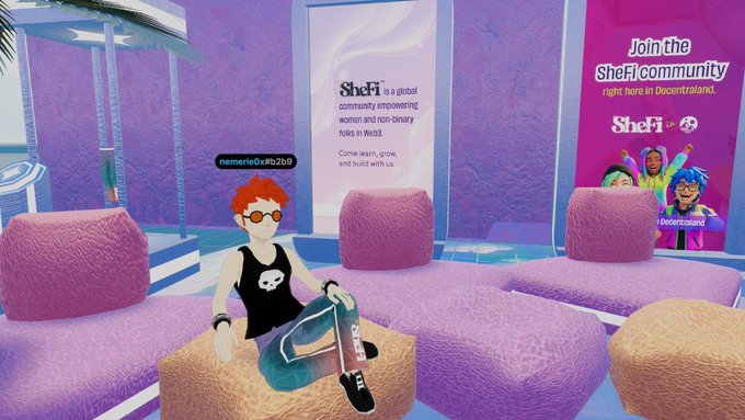 A red-haired avatar wearing sunglasses and a black tank top lounges on fluffy pink and orange cushions in a virtual booth promoting "SheFi," a global community empowering women and non-binary folks in Web3, alongside a "Join the SheFi community right here in Decentraland" banner.