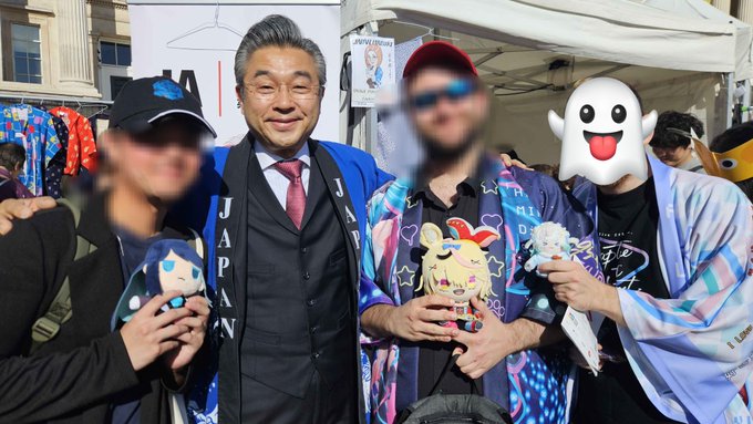 A group photo at what appears to be a convention shows several people, including an older man in a blue 'JAPAN' jacket, holding plushies, with one person's face obscured by an emoji.