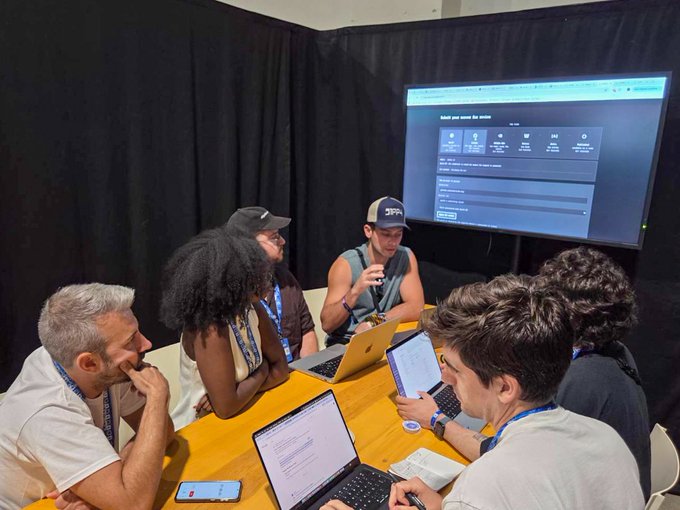 Group of five people seated around a wooden table working on laptops, with one man gesturing while speaking; a large screen displays a webpage in the background.