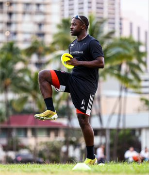Photo of a muscular soccer player in a black kit holding a yellow training ball, performing a high-knee drill on a grass field with palm trees and city buildings in the background.