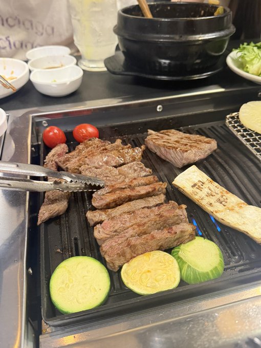 A sizzling Korean BBQ grill features marinated beef strips, cherry tomatoes, zucchini slices, and king oyster mushroom cooking alongside various side dishes and sauces. The image visually captures the "super delicious Korean beef" mentioned in the caption, showcasing the meal experience at the @MaraCakeHotSale dinner event.