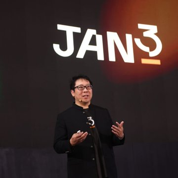 A man in a black suit and glasses speaking on stage in front of a large screen displaying the text 'JAN3'.