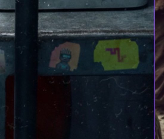 Graffiti-style artwork on a dark surface showing two head silhouettes: a figure with spiky hair in orange/pink on the left, and a lizard-like character in yellow with text resembling 'ZL' on the right, viewed through a wet window.