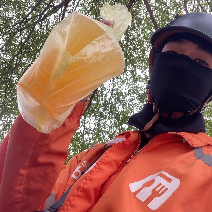 A delivery driver wearing an orange jacket and black helmet/face mask holding up a plastic bag containing an orange/yellow food item, photographed from below against trees.