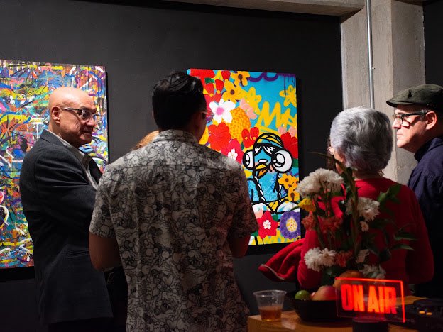 Several people in a gallery viewing colorful pop-art style paintings, with an 'ON AIR' sign visible in the foreground.