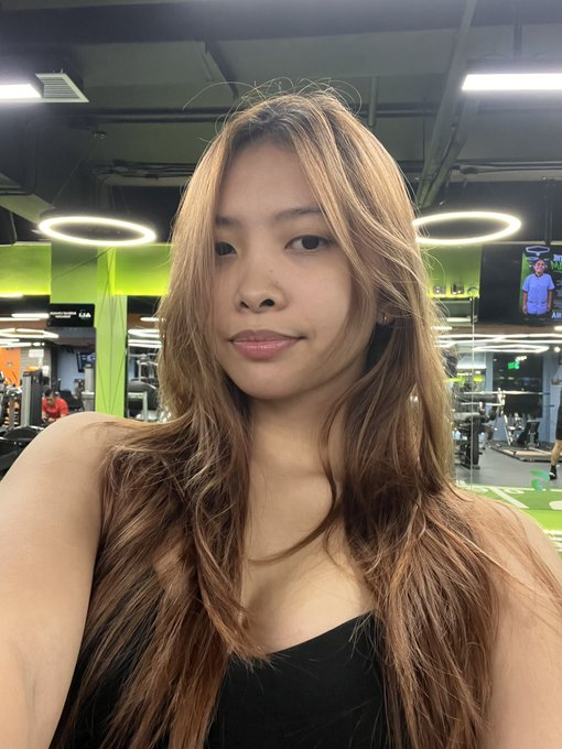 A selfie of a young woman with long light brown hair wearing a black top, taken in a gym with distinctive ring-shaped lights visible in the background.