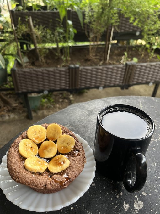 A breakfast scene featuring a chocolate pancake topped with banana slices and syrup on a white plate, served with a black mug of coffee on an outdoor table.