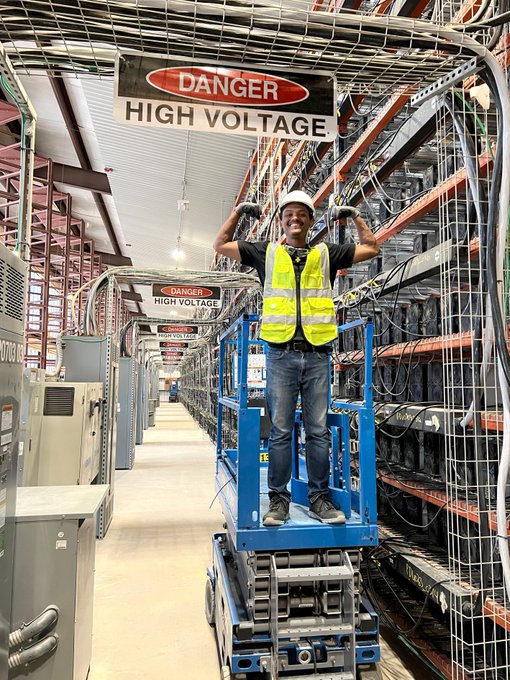 A smiling worker in a hard hat and yellow safety vest flexes his biceps while standing on a blue scissor lift between rows of mining equipment, beneath prominent "DANGER HIGH VOLTAGE" signs, illustrating the behind-the-scenes workforce that keeps bitcoin mining facilities running.
