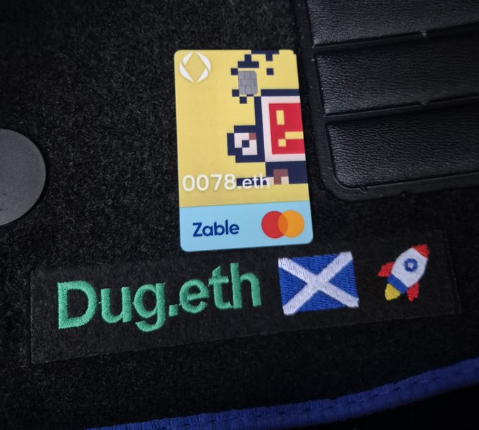 A yellow Zable Mastercard debit card labeled '0078.eth' sitting on a black wallet next to a patch reading 'Dug.eth' with a Scottish flag and rocket ship embroidered.