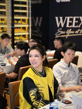 A woman in a yellow and black sports jersey sitting at a table at what appears to be a WEEX event, with other attendees visible in the background.