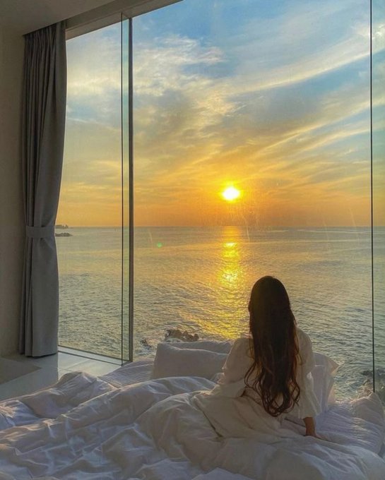 A woman with long dark hair sits on a bed in a white robe, looking out a large floor-to-ceiling window at a sunset over the ocean.