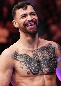 A muscular fighter with a chest tattoo and a blue mouthguard grins confidently after his victory in the octagon, contrasting with the betting news about Arman Tsarukyan, who is the subject of a $23,000 wager on Polymarket.