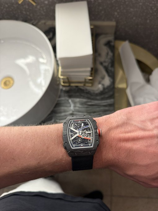 A wrist wearing a black carbon-fiber skeleton watch with an orange second hand, positioned above a marble bathroom counter featuring a white sink, a gold-accented tissue holder, and a brass fixture.