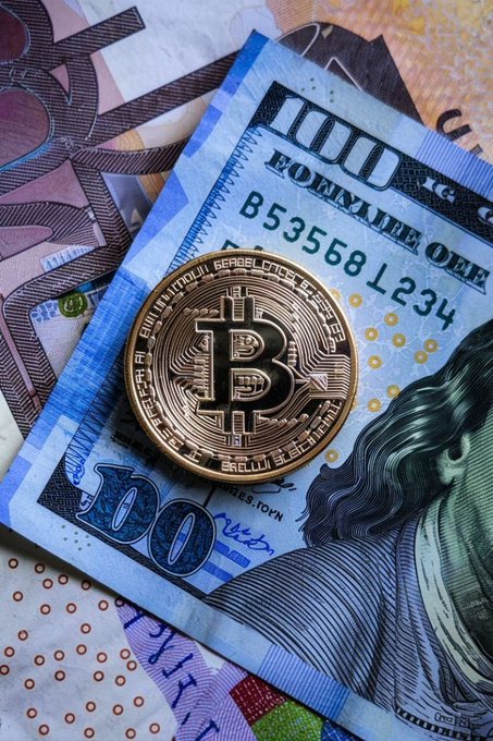 A close-up shows a physical Bitcoin coin resting on top of various paper currencies, including a 100 hryvnia note.