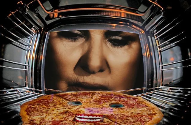 A woman's face peers intensely into an open oven at a pizza that has googly eyes and pink lips drawn on it, giving it a cartoonish face.