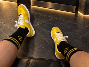 A pair of yellow sneakers paired with black and yellow Binance-branded socks rests on a dark surface, complementing the post's promotion of a Binance Mid-Autumn Festival gift box giveaway featuring the same shoe style.