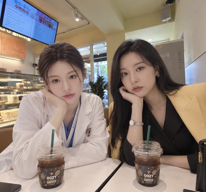Two young women posing together at a Starbucks inside what looks like a BBK (베이커리) café, with menu boards visible in the background and iced coffee cups branded 'OOZY' on the table.