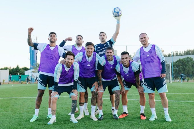 A group of seven men, including Lionel Messi, pose together on a soccer field wearing purple and white Argentina training gear with Coca-Cola and Binance logos.